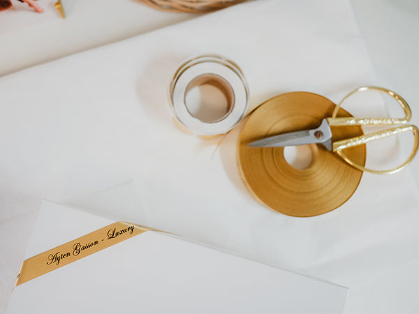 Gold ribbon with scissors on a white surface, featuring the brand 'Ayten Gasson'.