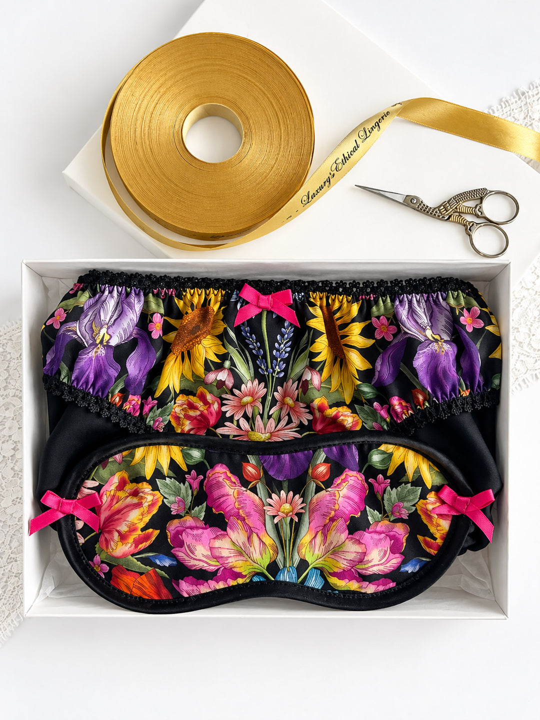 A gift set containing a black satin sleep mask and matching underwear set with a vibrant multicolor floral print, pink bows, and black lace trim, presented in a white box alongside a roll of gold ribbon and silver scissors.