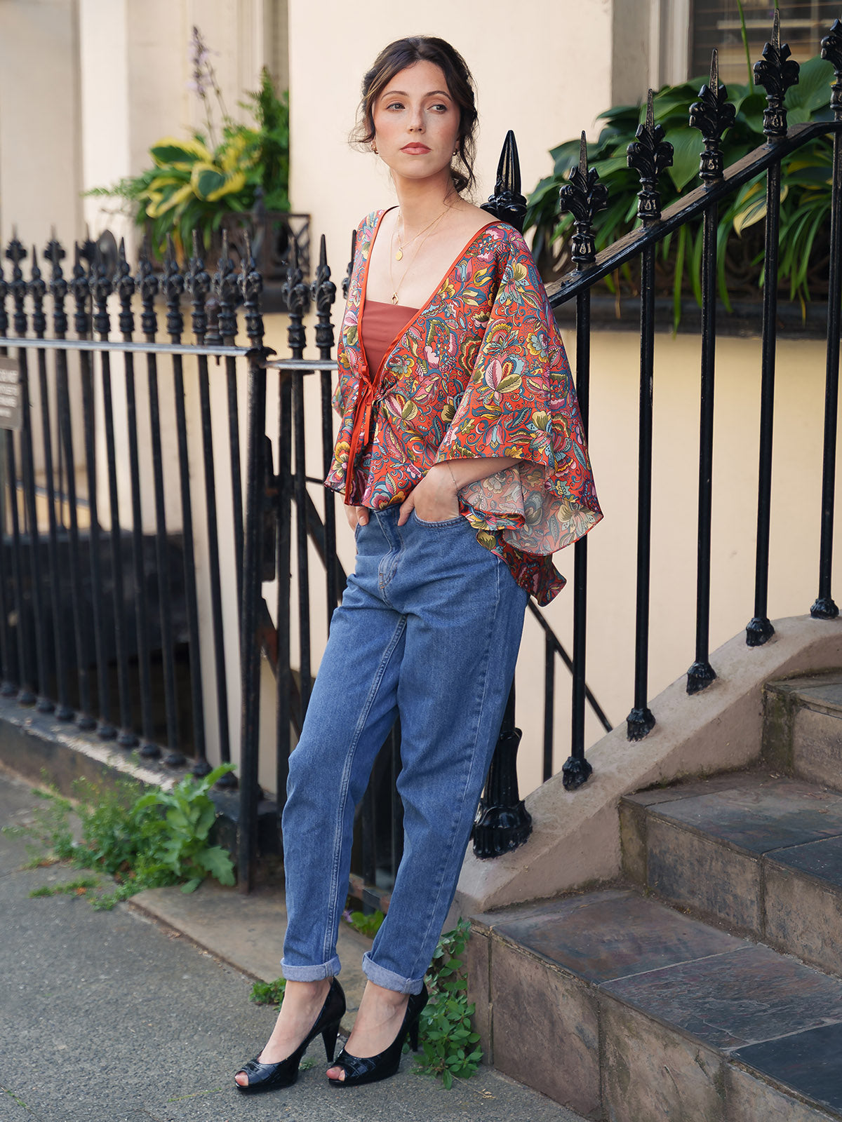 Woman wearing a colorful patterned kimono over a red top and blue jeans, standing on steps with a black metal railing and greenery in the background.