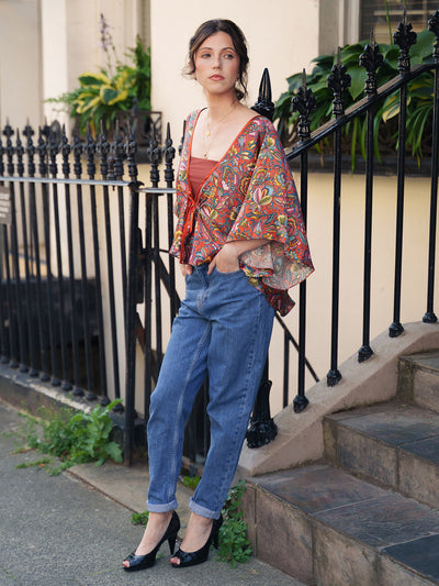 Woman wearing a colorful patterned kimono over a red top and blue jeans, standing on steps with a black metal railing and greenery in the background.