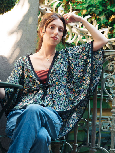Woman wearing a floral Rachel Meadow Liberty Silk Bed Jacket kimono-style garment sitting on a bench outdoors.