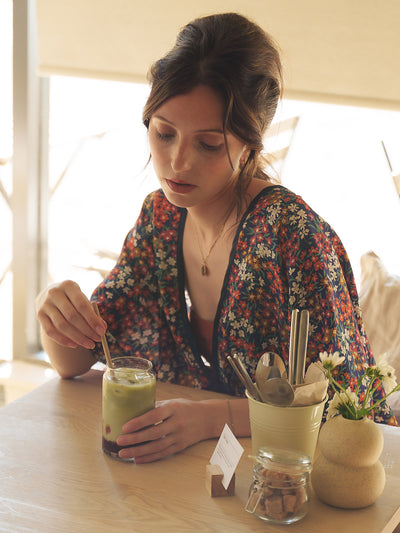 Woman sitting at a table with a green smoothie, wearing the Sea Blossom Liberty Navy Silk Bed Jacket by Ayten Gasson