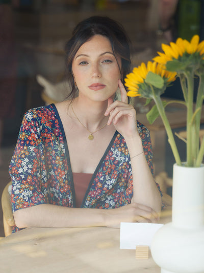 Woman in a Sea Blossom Liberty Navy Silk Bed Jacket sitting at a table with sunflowers in the background